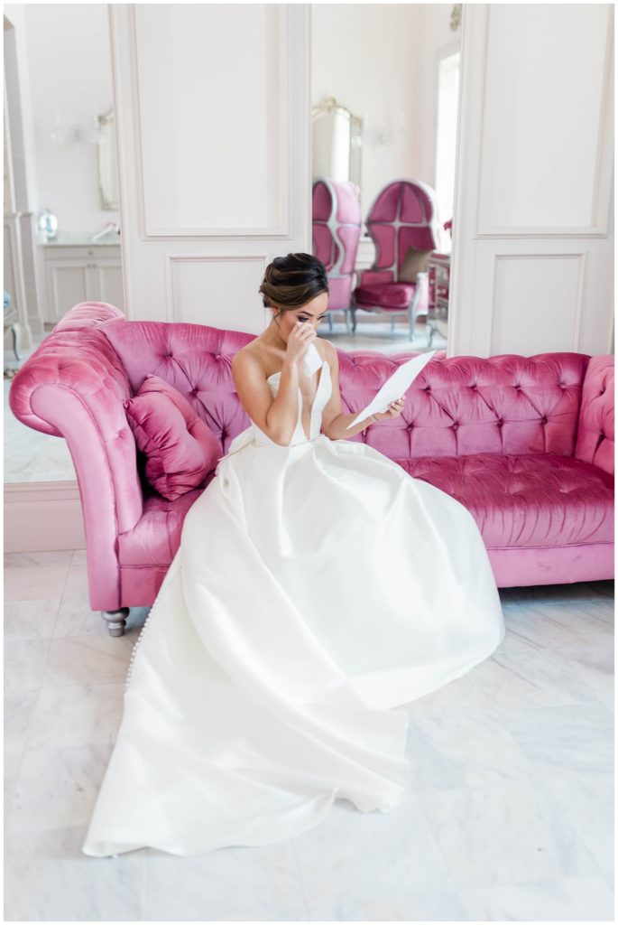 bride reading a letter from her groom as she sits in a pink couch in her bridal suite