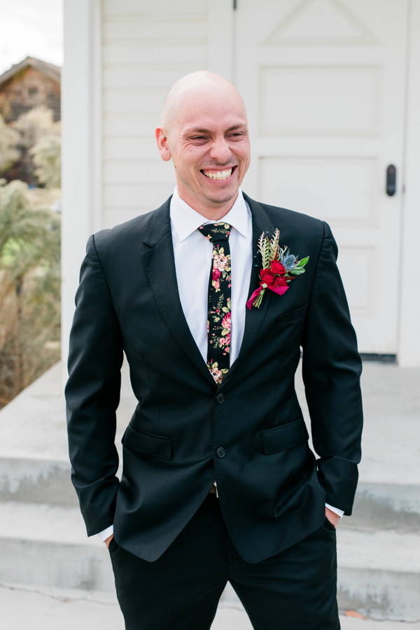 groom smiling with his hands in his pockets with a red boutonniere 