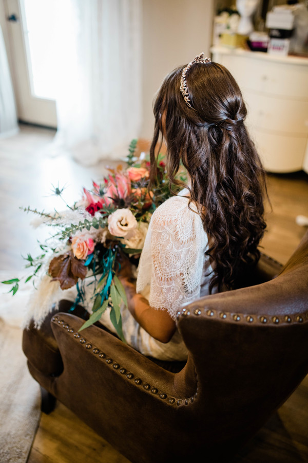 bride sitting on a brown leather chair while holding her wedding flowers with the image taken from behind to highlight her bridal hairstyle
