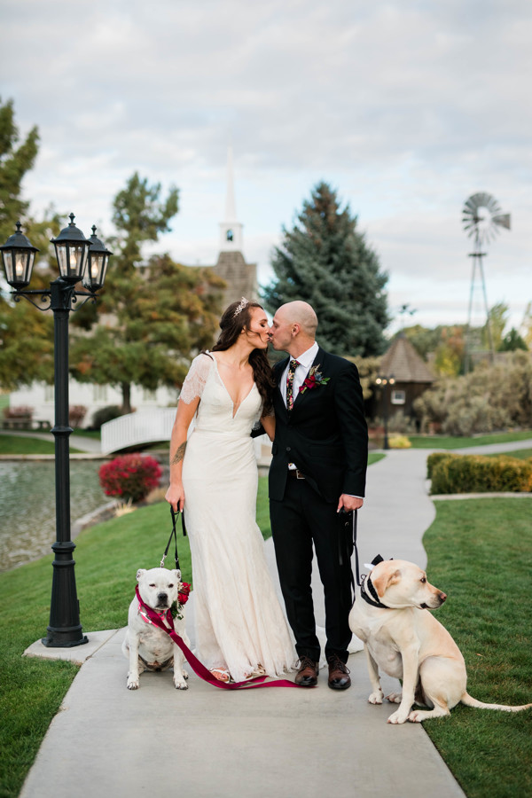bride and groom kiss while holding dogs Lucy and McKoy with floral collars on wedding day