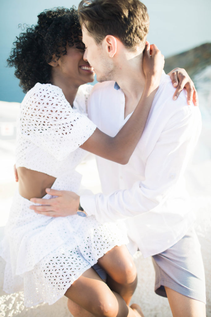 woman sitting on mans lap and holding his jaw line as she leans in to kiss him during their destination Greek engagement session