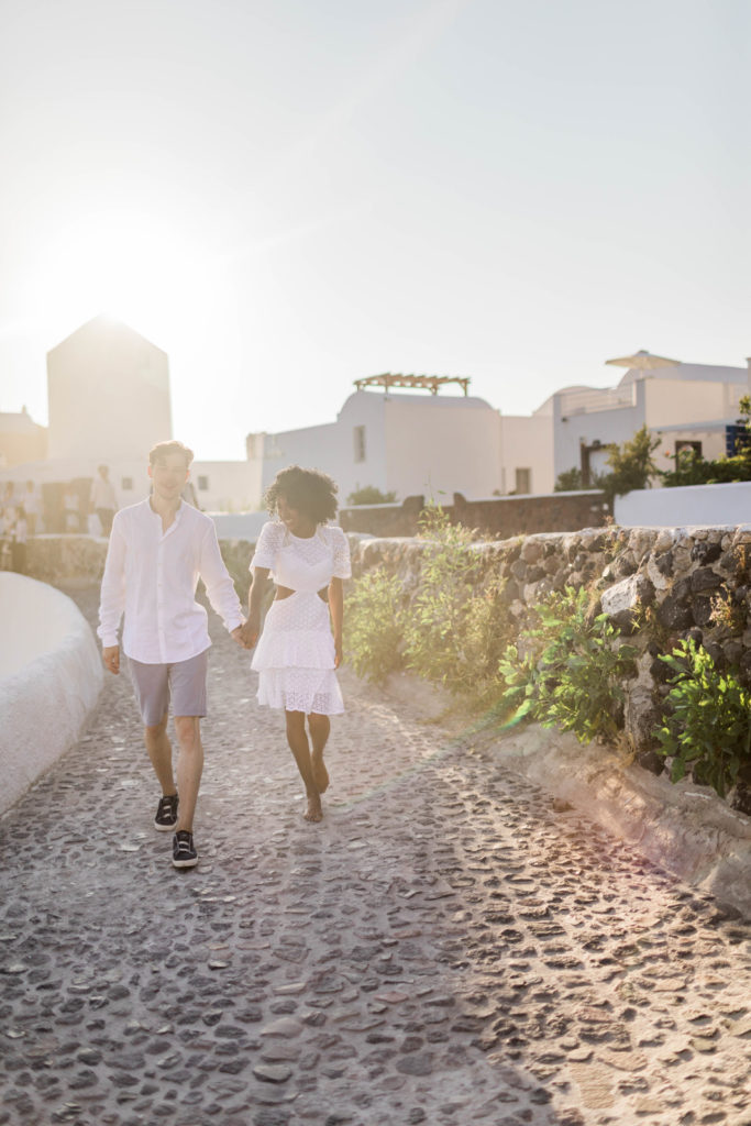 man and woman walking on a cobblestone path in Greece while holding hands with the sun setting behind them