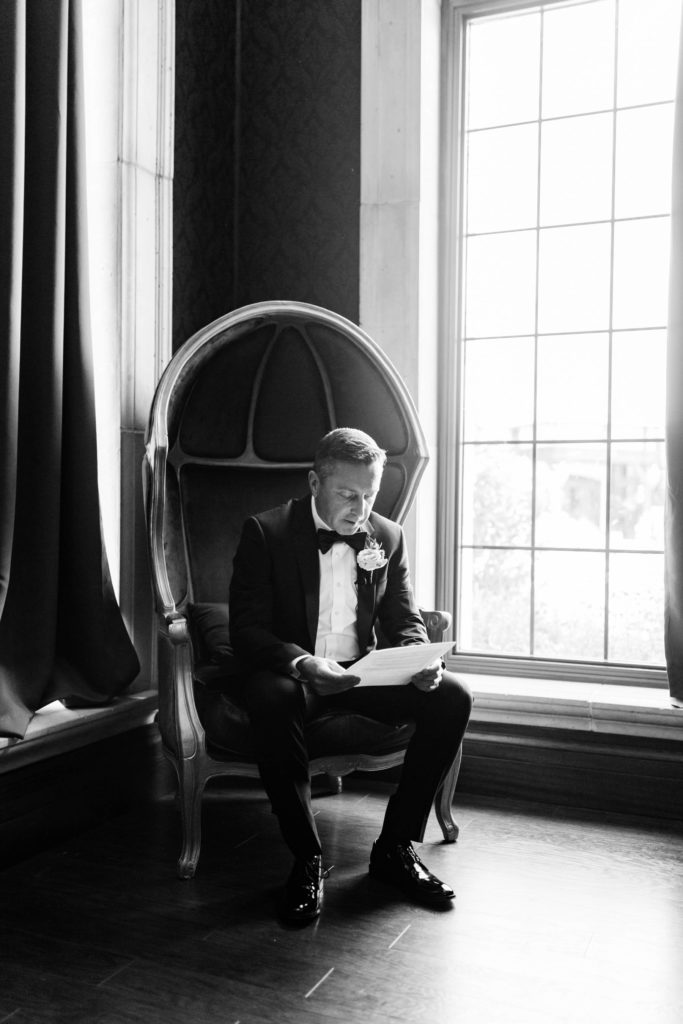 groom sitting in a wicker chair and reading a letter from his bride near a window