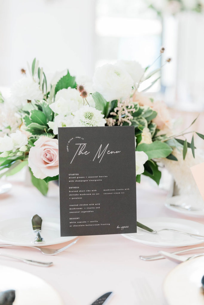 wedding invitation on a black canvas with silver lettering against a floral center piece on a wedding table
