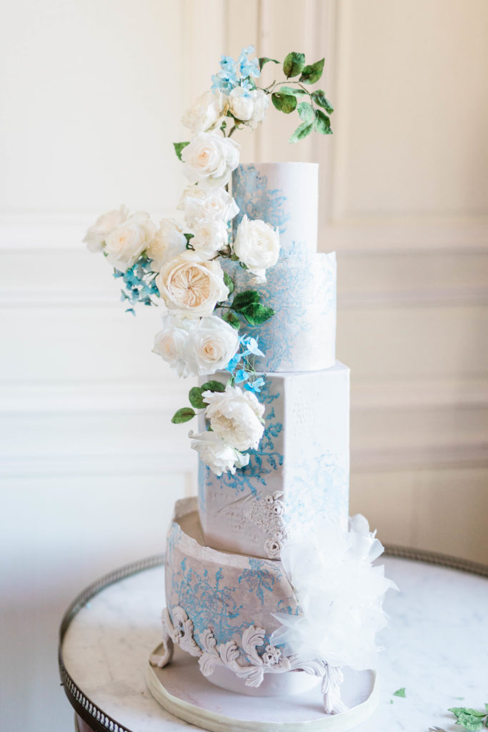 four tier wedding cake with a variety of shapes has a large white rose floral arrangement on the side of the cake