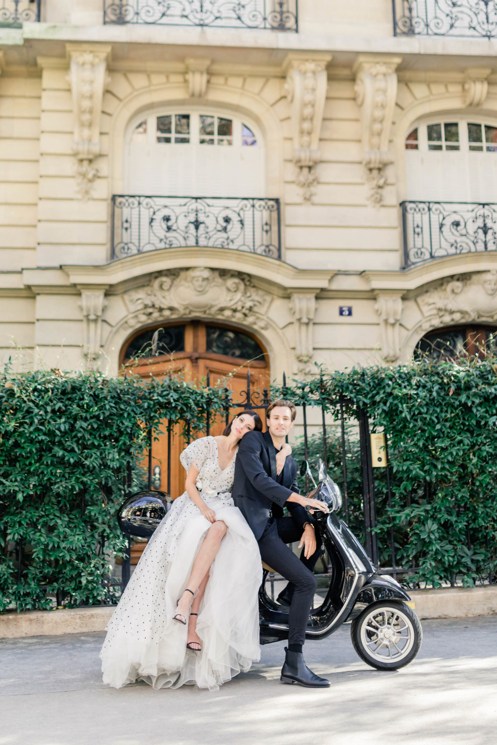 man and woman sitting together on a motorcycle in front of a black iron fence with ivy growing on it in France for their destination engagement 