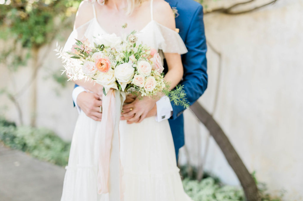 bride and groom embracing one another in a courtyard with the man behind his bride as she holds her wedding flowers