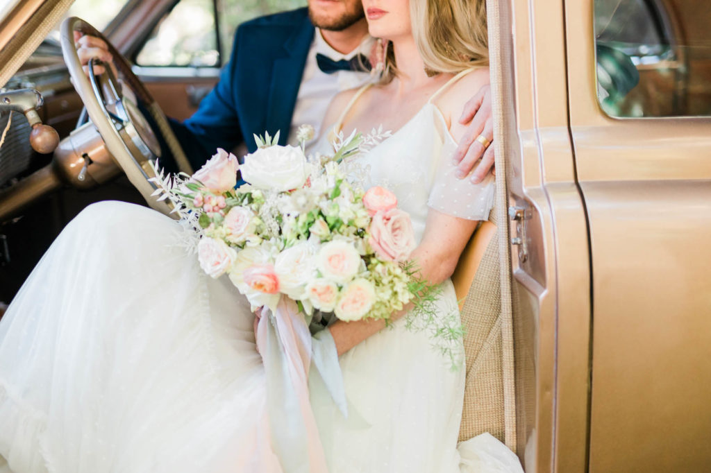 bride and groom sitting in a brown vintage car with the bride holding a beautiful white and pink bouquet