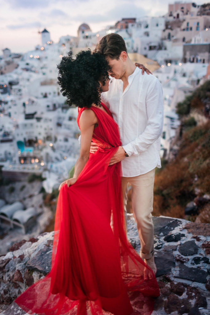 man and woman holding each other as their noses touch for a romantic engagement photo pose with the man holding to womans waist as they stand on a cliff with Greece's skyline behind them 