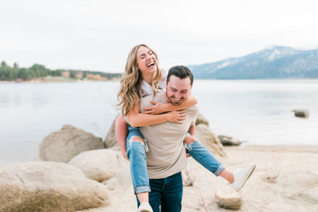 woman piggy backing on the man as they walk on the rocks of a beach and the man an woman laugh together