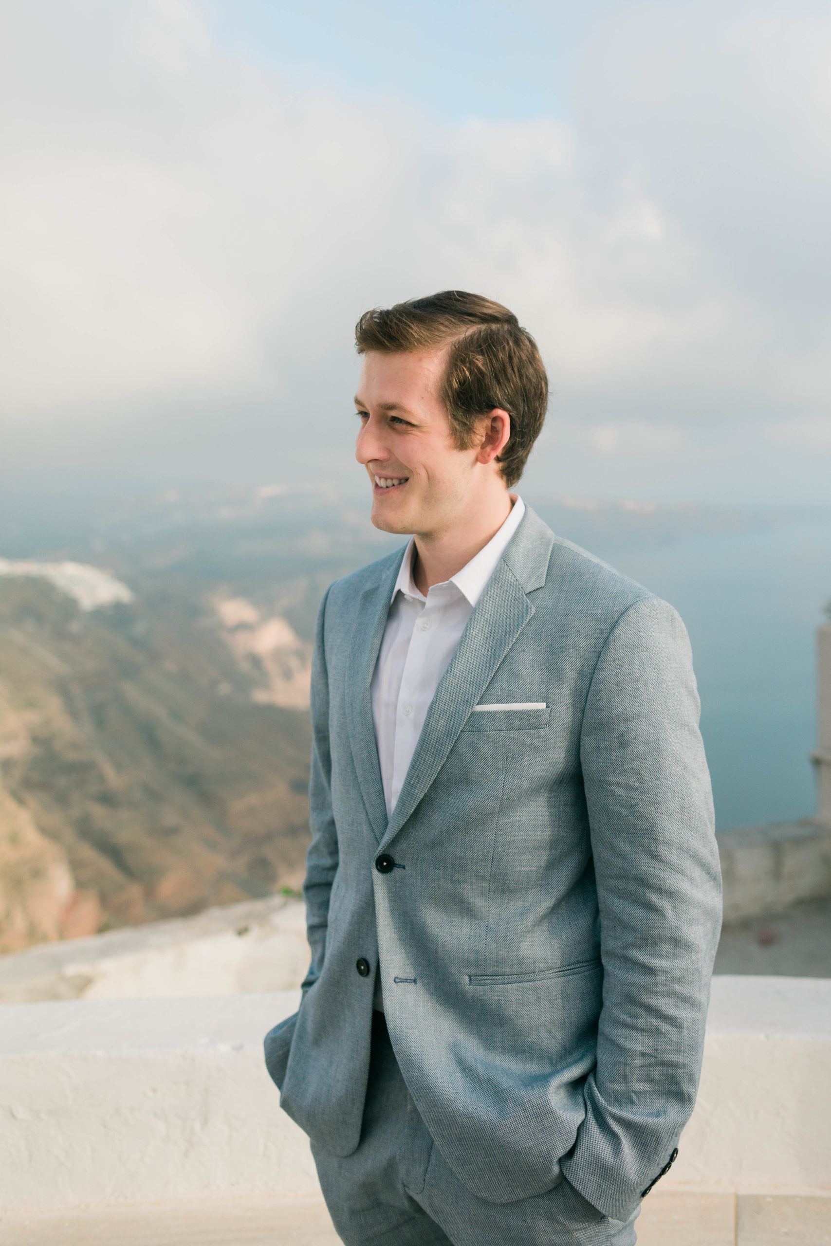 groom looking off to the distance and he smiles at his bride with the ocean behind him as he wears a grey suit