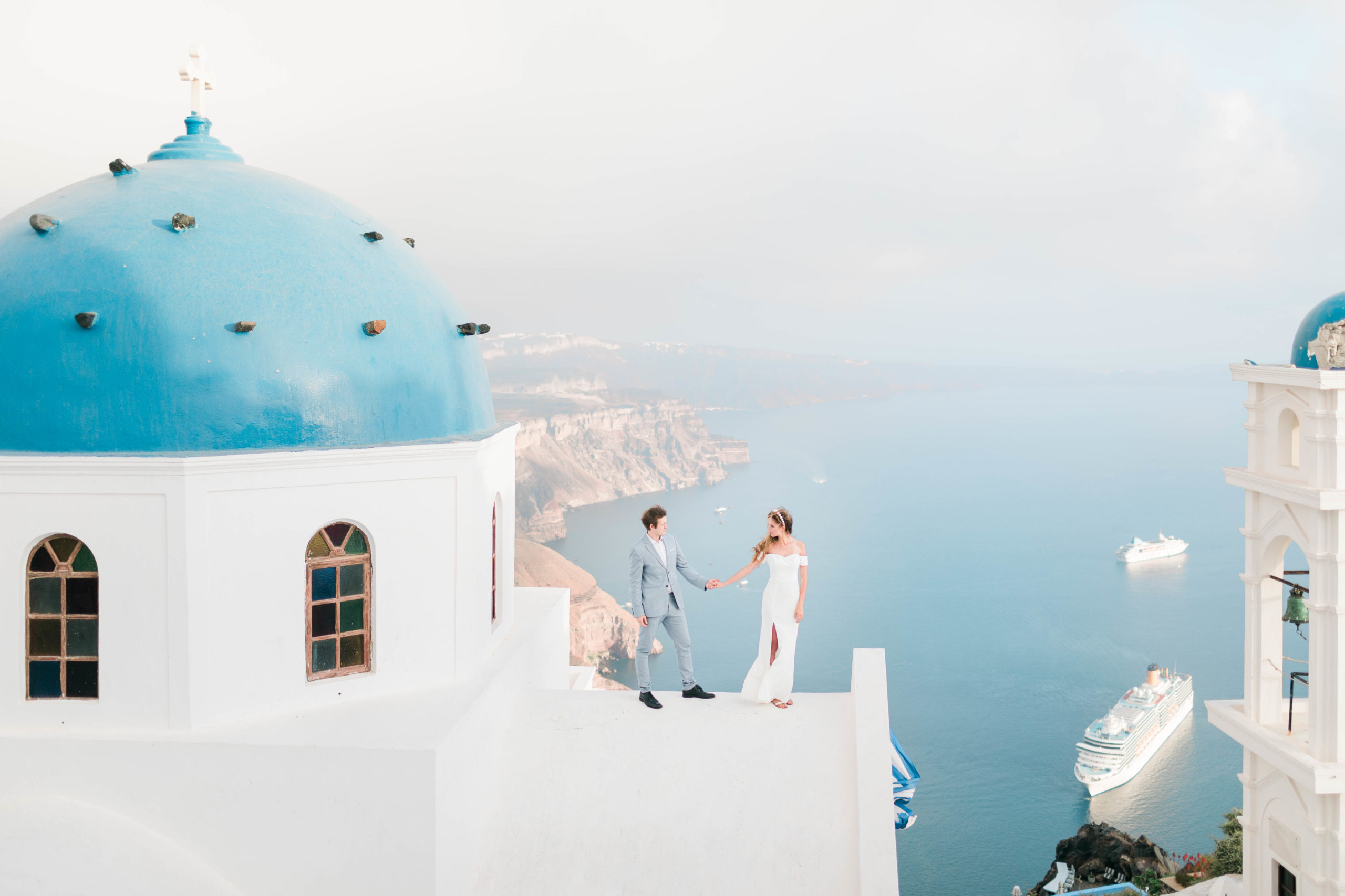 bride and groom holding hands as they stand together on a wall of a white building in Greece with the ocean behind and below them
