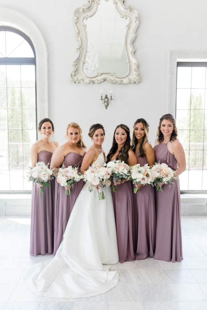 bridesmaids in light purple pink dresses stand with the bride in the bridal suite while holding white bouquet and smiling at the Boise wedding photographer 
