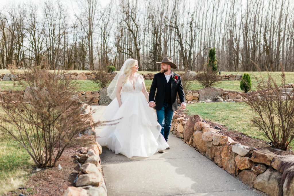 bride adn groom holding hands as the bride wears a ruffled skirt wedding dress and the groom is in jeans and a blazer with a cowboy hat on top of his head at luxury Boise wedding 