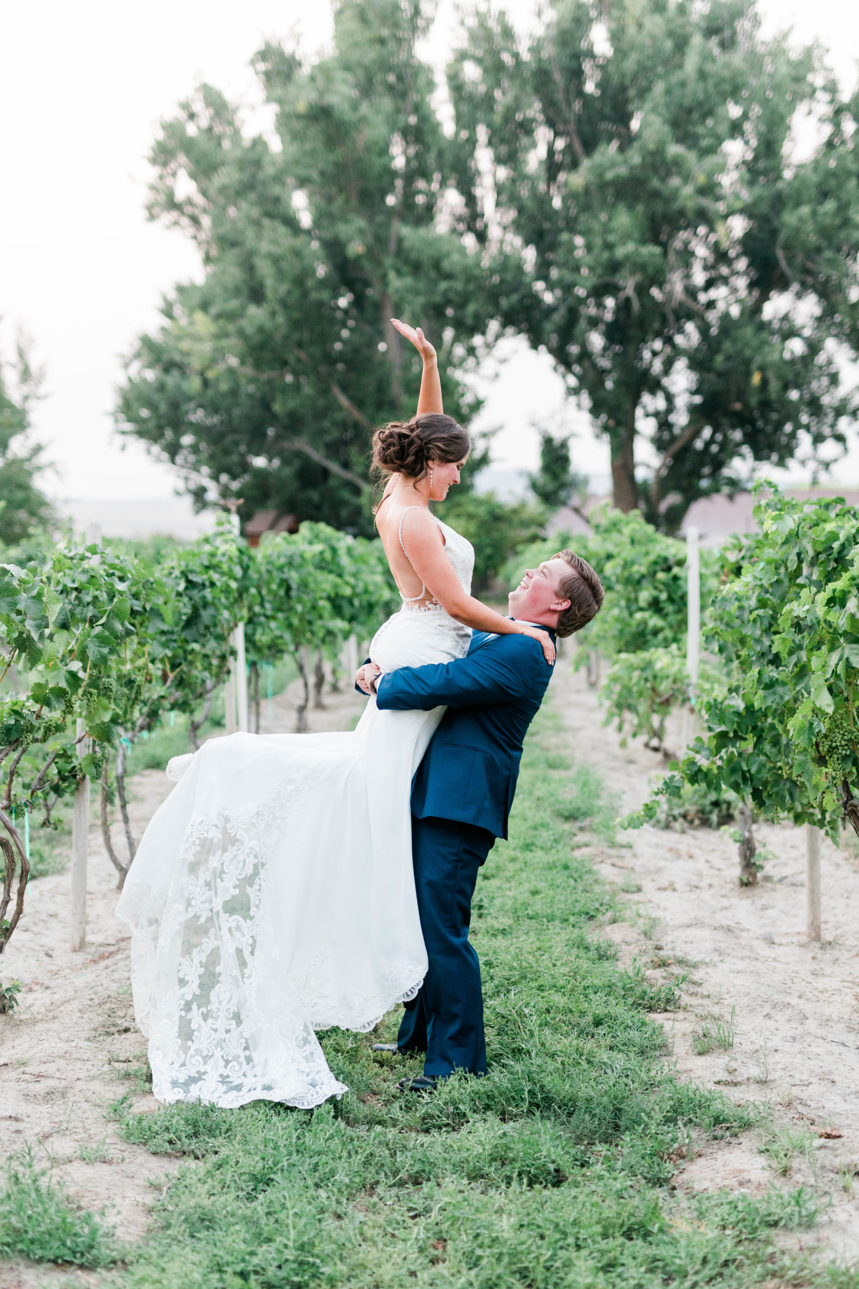 Boise vineyard wedding venue with groom picking up his bride from under her bottom as the bride throws a hand into the air