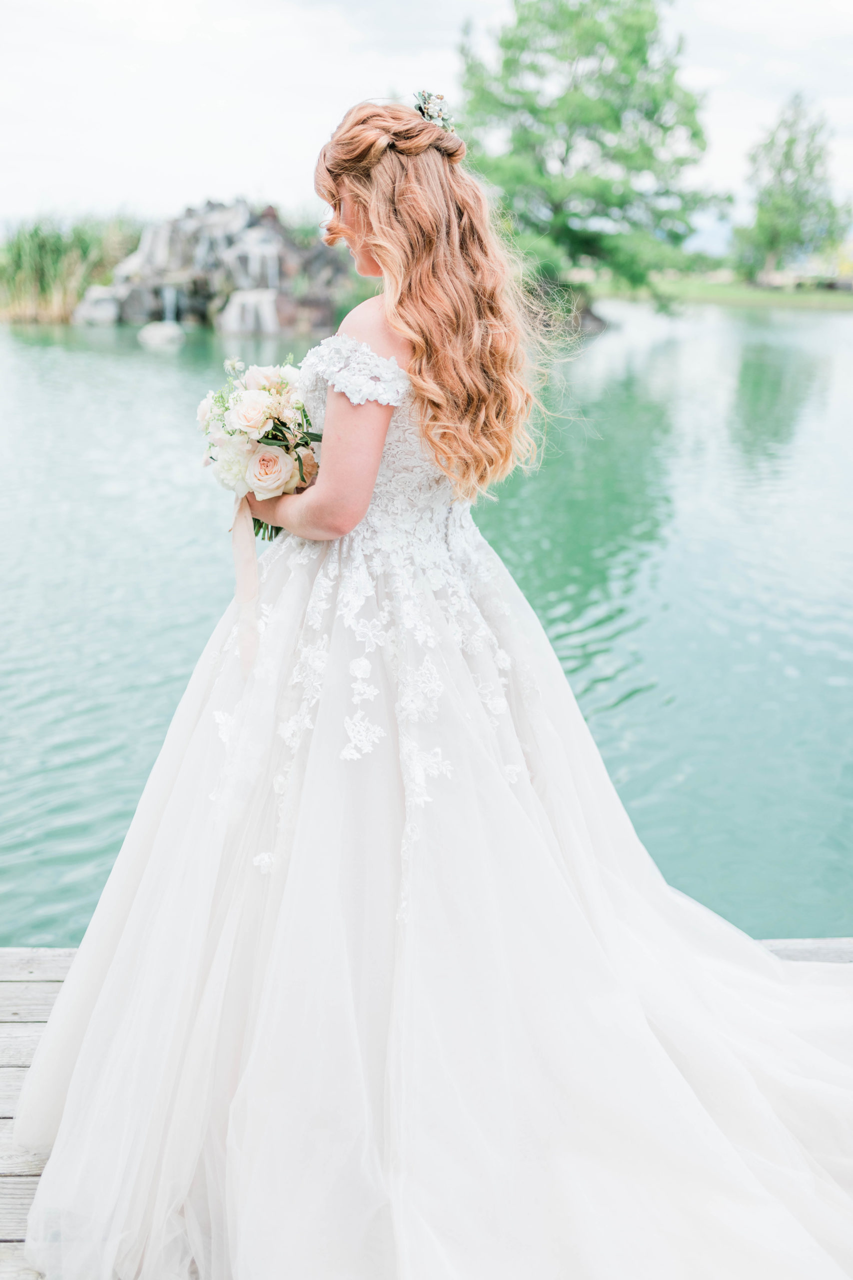 bride in a florals lace wedding gown with an off the shoulder sleeve holds her bouquet and looks out into a lake