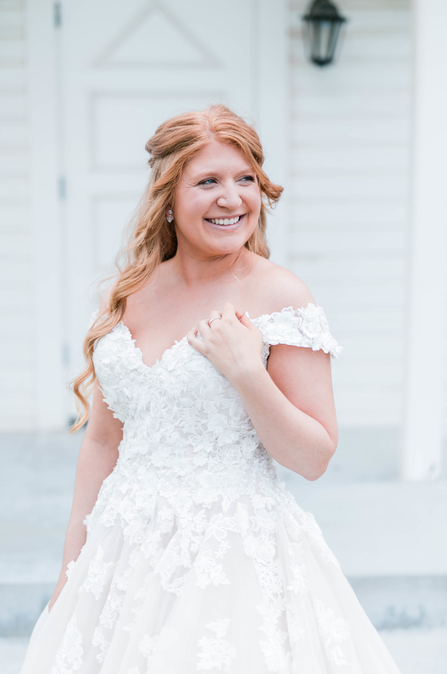 bride in a floral lace wedding dress with off the shoulder sleeves holds her hair and looks to the side while smiling