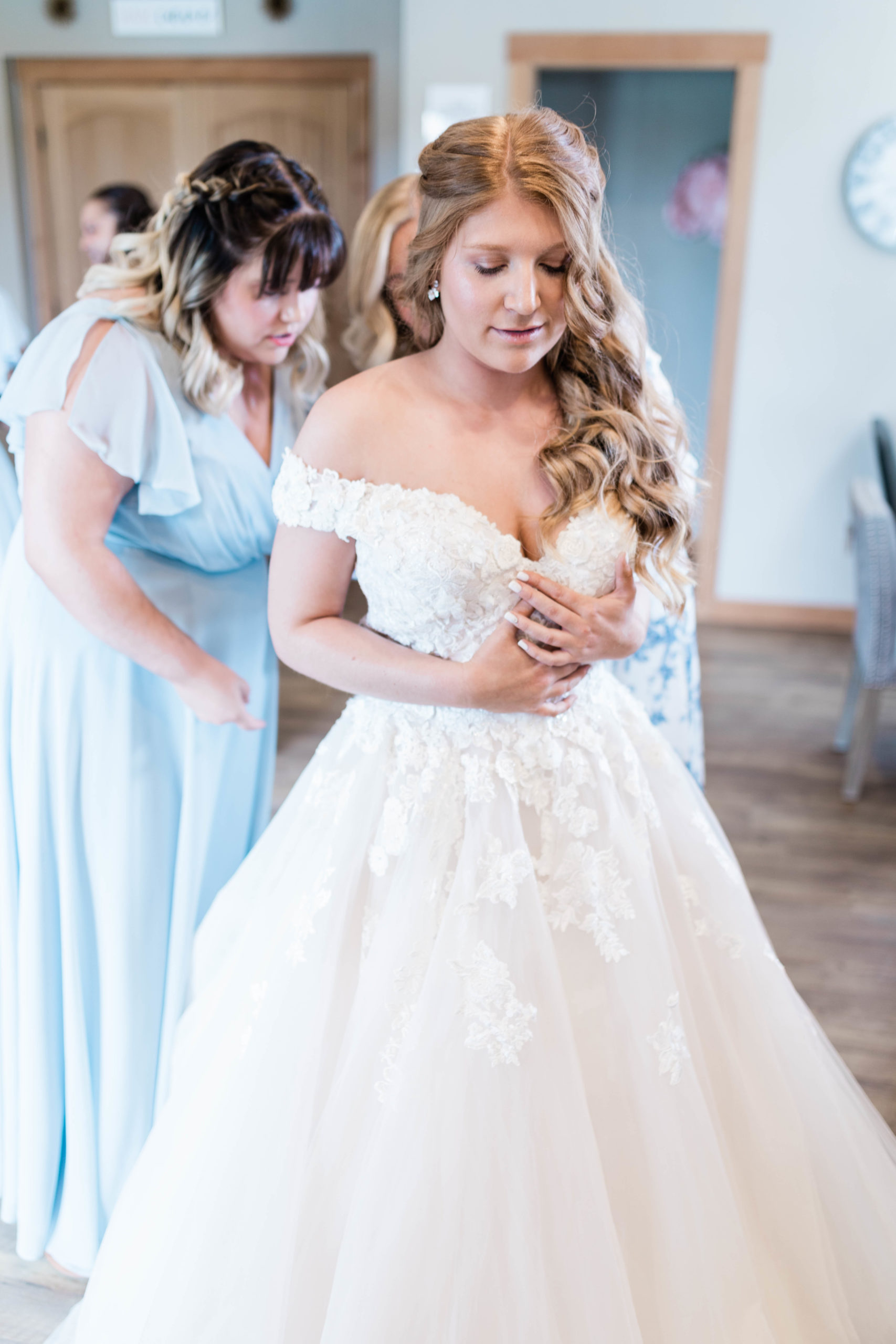 bride holding her hands together as her mother buttons the back of her wedding gown with her bridesmaids