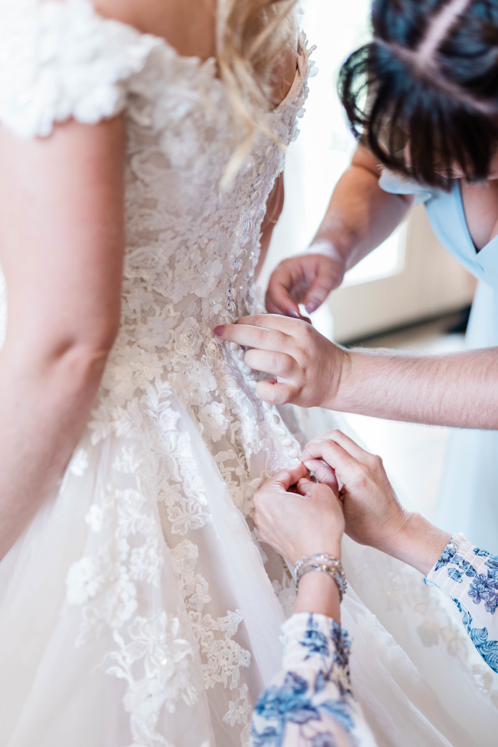 mother of the bride and maid of honor helping the bride by buttoning the back of her floral lace wedding gown in the bridal room in Still Water Hallow wedding venue