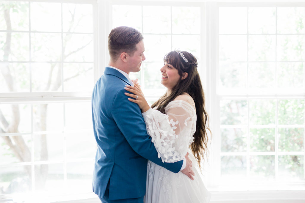 bride and groom in a white room with large bay windows as they dance together before their wedding ceremony