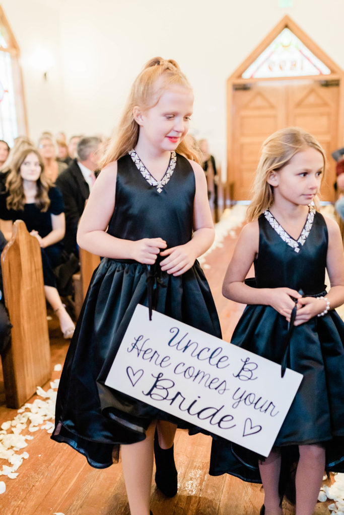 Boise wedding photographer captures flower girls walking down aisle holding sign 'here comes the bride'